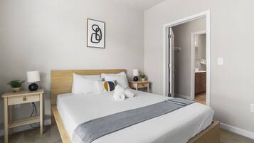 A neatly made bed with a wooden headboard, two bedside tables with lamps, a small plant, and a framed abstract artwork on the wall.