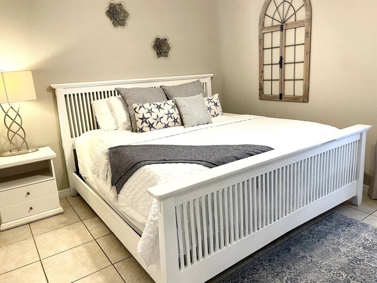 A neatly made bed with a white headboard and footboard, a bedside table with a lamp, and a window with a wooden frame.