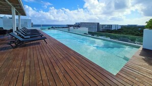 Una piscina infinita en la azotea con cubierta de madera y sillones de descanso, con vistas a la ciudad y al océano.