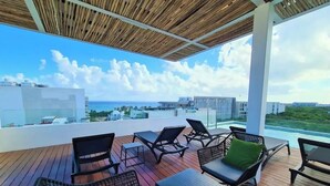 Terraza en la azotea con mobiliario exterior, vista al océano y edificios modernos al fondo.