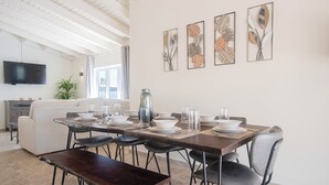 A dining area with a long table set for four, surrounded by black chairs, a wooden bench, and a large vase. A flat-screen TV is mounted on the wall above the sofa in the background.