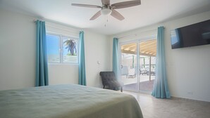 A bedroom with a bed, a ceiling fan, a TV, and a view of an outdoor patio with a chair and curtains.