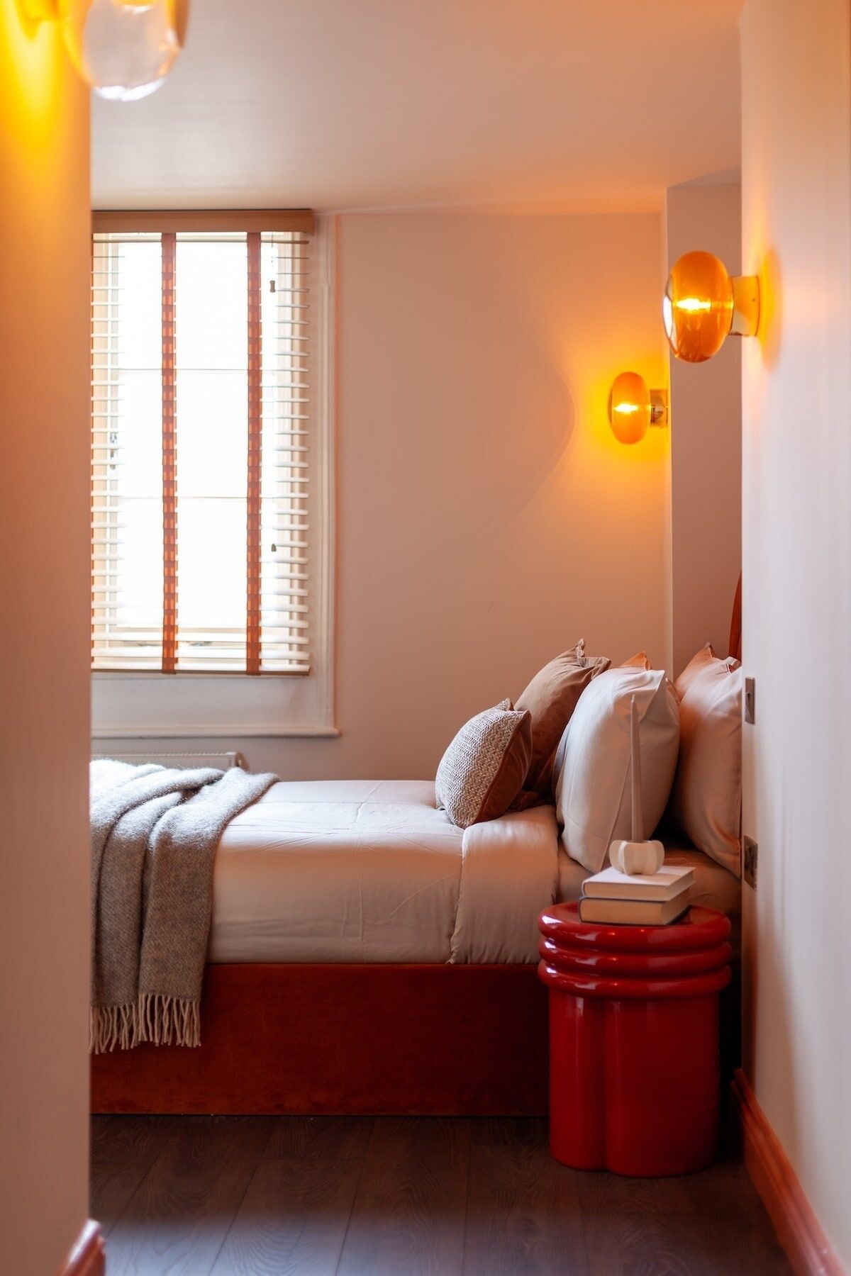 A cozy bedroom with a bed, pillows, a bedside table with books, and a window with blinds.