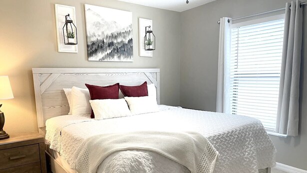 A neatly made bed with white bedding and a few red pillows, a bedside table with a lamp, and framed artwork on the wall. - Spotless Escape in the City`s Core 837 (Clermont)