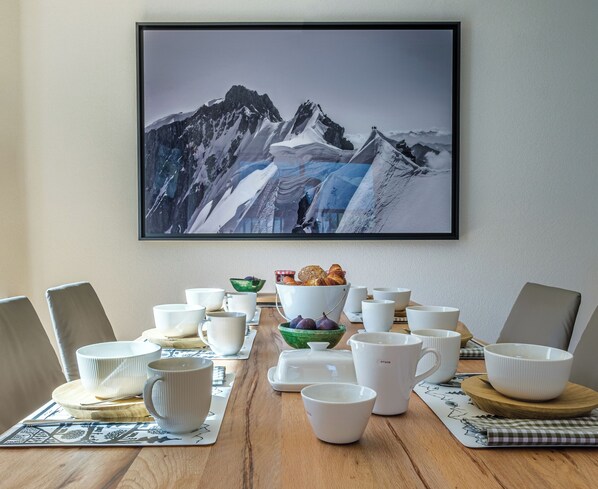Una mesa de comedor con tazas y cuencos de cerámica blanca, y una hogaza de pan, frente a un cuadro enmarcado con un paisaje de montaña.