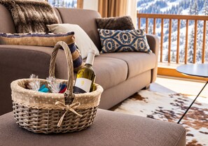 A wicker basket with a wine bottle and items on a cushioned surface in a living room with a view of snow-covered mountains.