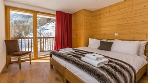Une chambre avec un grand lit, une tête de lit en bois, une chaise et un balcon agrémenté d’un rideau rouge.