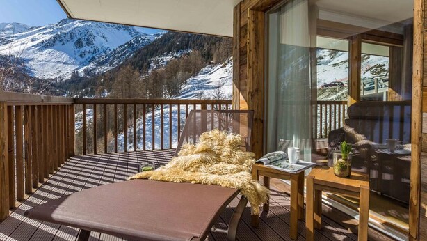 Une terrasse en bois offrant une vue sur des montagnes enneigées, une chaise rembourrée, une petite table avec une plante et une cloison en verre.