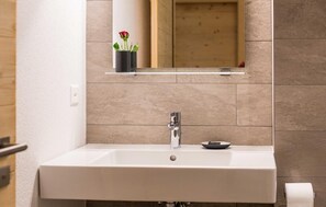 Un baño con un lavamanos blanco, un espejo encima y un pequeño florero con una flor.