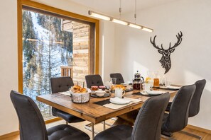 A dining area with a wooden table, black chairs, and a view of trees through a large window.