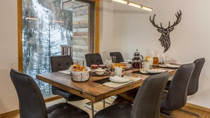 A dining area with a wooden table set for four, surrounded by chairs, with a large window offering a view of a forest. - Luxury Apartment, 3 Bedrooms with Jacuzzi Chalet Rahas A Grimentz - By Mrs Miggins (Grimentz)