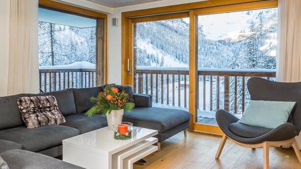 Ett mysigt vardagsrum med en grå soffa, ett vitt soffbord och en grå fåtölj. Det finns ett stort fönster med utsikt över ett snötäckt berglandskap.
