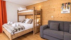 En våningssäng med kuddar och ett täcke, en grå soffa, träväggar och en skylt med texten ”VALAIS, solens land”.