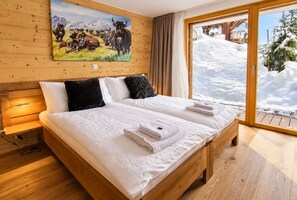 Una cama con estructura de madera, ropa de cama y toallas blancas, un cuadro de ganado en la pared y vistas a un paisaje nevado a través de un ventanal.