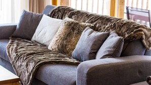 Ein graues Sofa mit einem Fellüberwurf und drei Kissen in einem Raum mit Holzbalkon und Fenster.