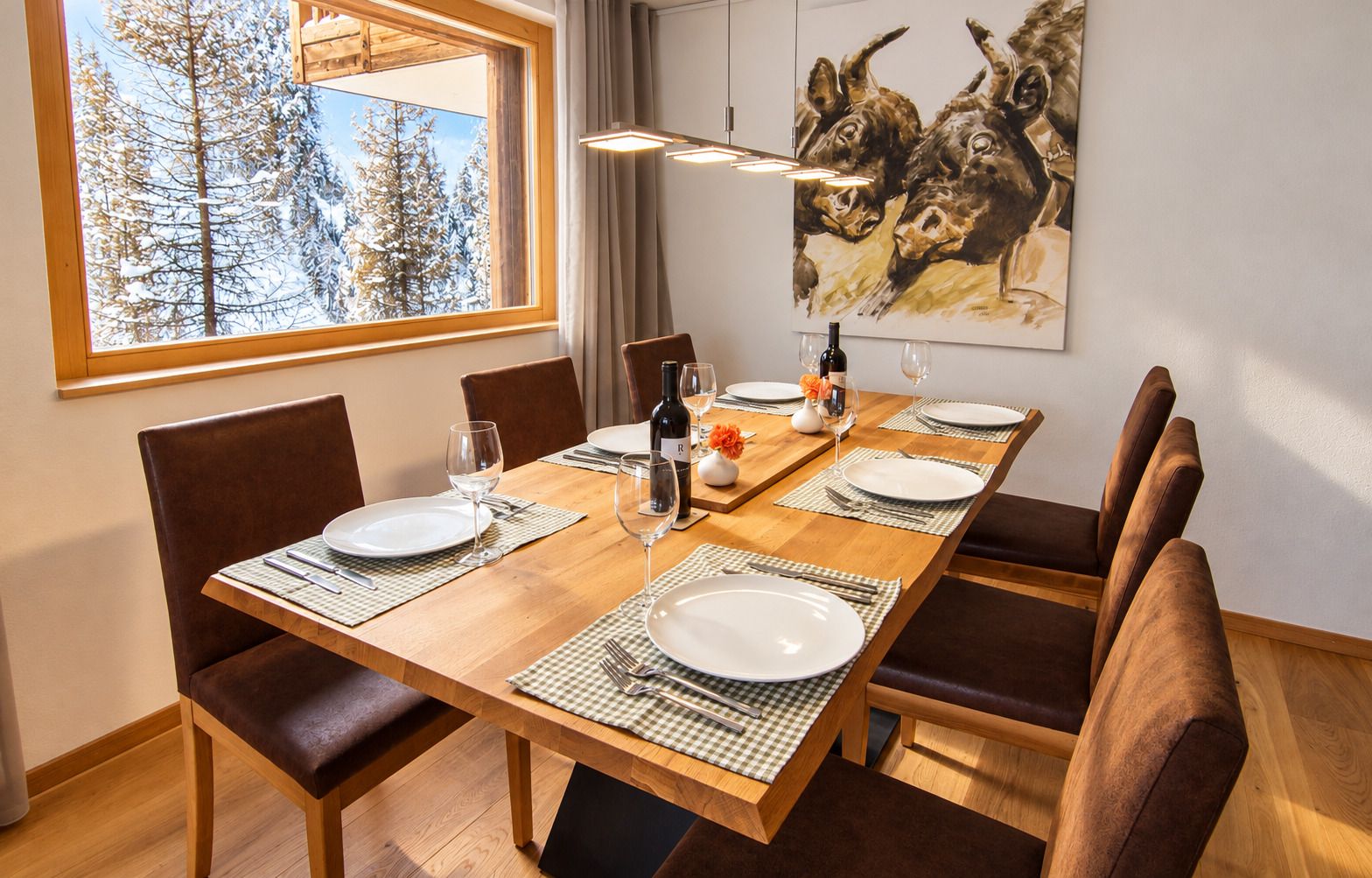 Ein Esszimmer mit einem Holztisch, gedeckt für vier Personen, Stühlen, einem Gemälde an der Wand und einem Fenster mit Blick auf schneebedeckte Bäume.