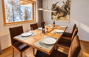 Un comedor con una mesa de madera dispuesta para cuatro, sillas, un cuadro en la pared y una ventana con vistas a árboles cubiertos de nieve.