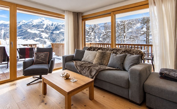 A cozy living room with a sofa, armchair, and coffee table. Large windows offer a view of snow-covered mountains.