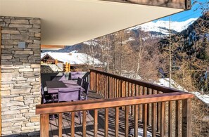 Een houten terras met een stenen muur, zitplaatsen buiten en uitzicht op besneeuwde bergen.