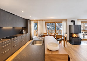 A modern kitchen with a central island, a dining area with a fireplace, and large windows with a view of trees.