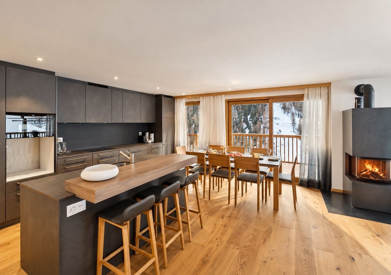 A modern kitchen with a central island, bar stools, a dining area with a view, and a fireplace.