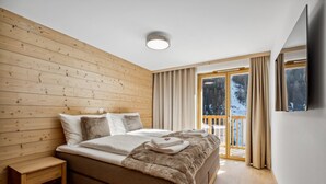 Een slaapkamer met een houten strookplafond, een bed met wit en bruin beddengoed, een houten bank en een groot raam met uitzicht op bomen.