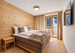 A bedroom with a wooden wall, a bed with a fur throw, a bedside table, and a view of a snowy landscape through a glass door.