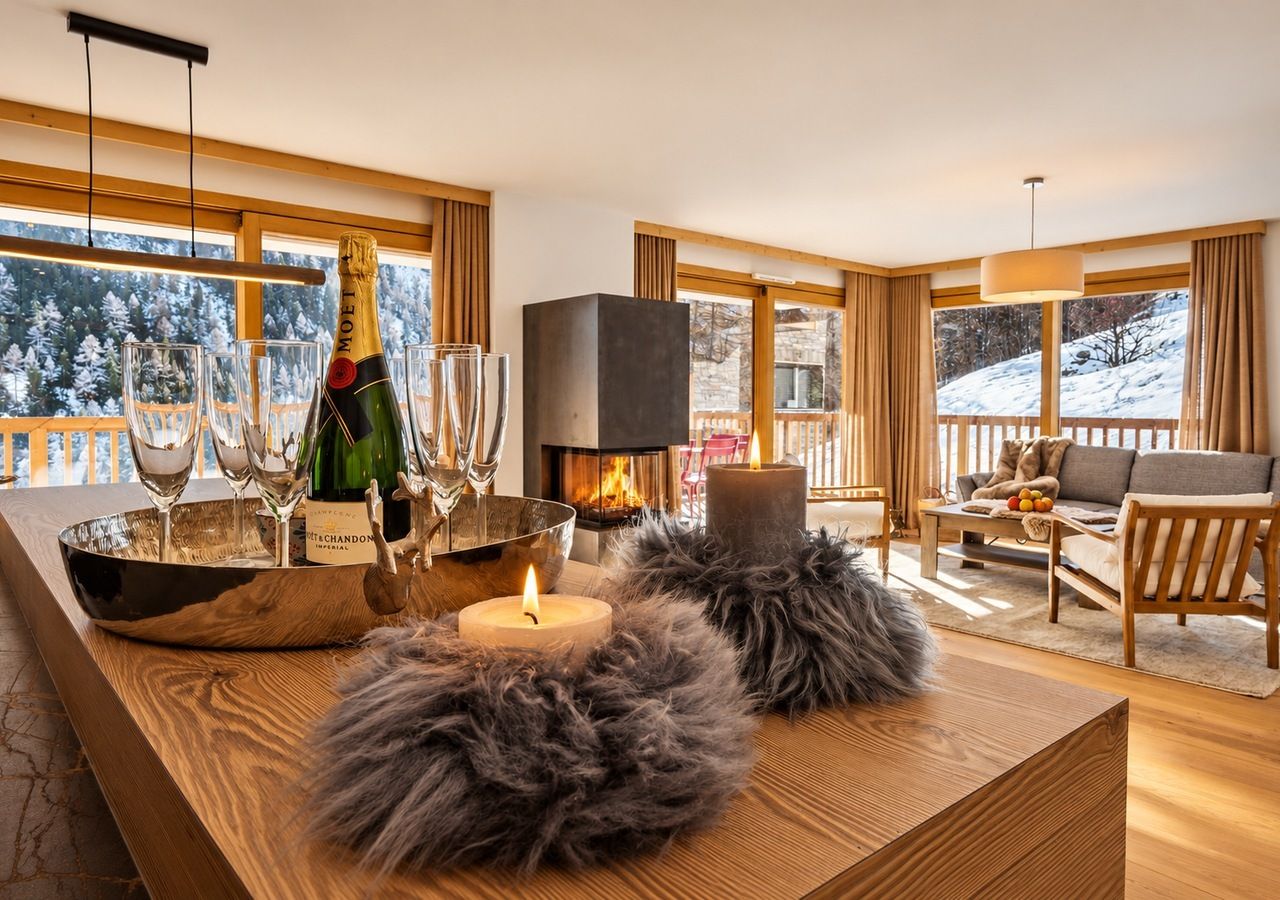 Ein gemütliches Wohnzimmer mit Kamin, einer Champagnerflasche, Gläsern und einer Kerze auf einem Holztisch.