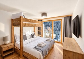 Een slaapkamer met een hemelbed, een nachtkastje met een lamp, een bankje en een groot raam met gordijnen.
