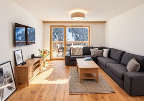 Ein modernes Wohnzimmer mit einem dunkelgrauen Sofa, einem Holztisch und einem großen Fenster mit Blick auf Bäume.