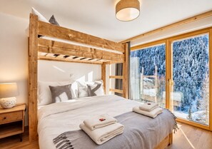 Ein Schlafzimmer mit hölzernem Kopfteil, einem Bett mit weißer Bettwäsche, einem Nachttisch mit Lampe und Blick auf verschneite Bäume durch große Fenster.