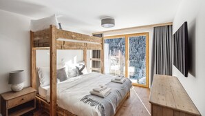 Un dormitorio con una cama litera de madera, una mesita de noche con una lámpara, un banco de madera y un ventanal con vistas a un paisaje nevado.
