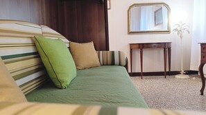 Una cama con ropa de cama a rayas, un almohadón verde y un almohadón marrón. Hay una mesita de noche de madera con un espejo y una pequeña mesa junto a la cama.