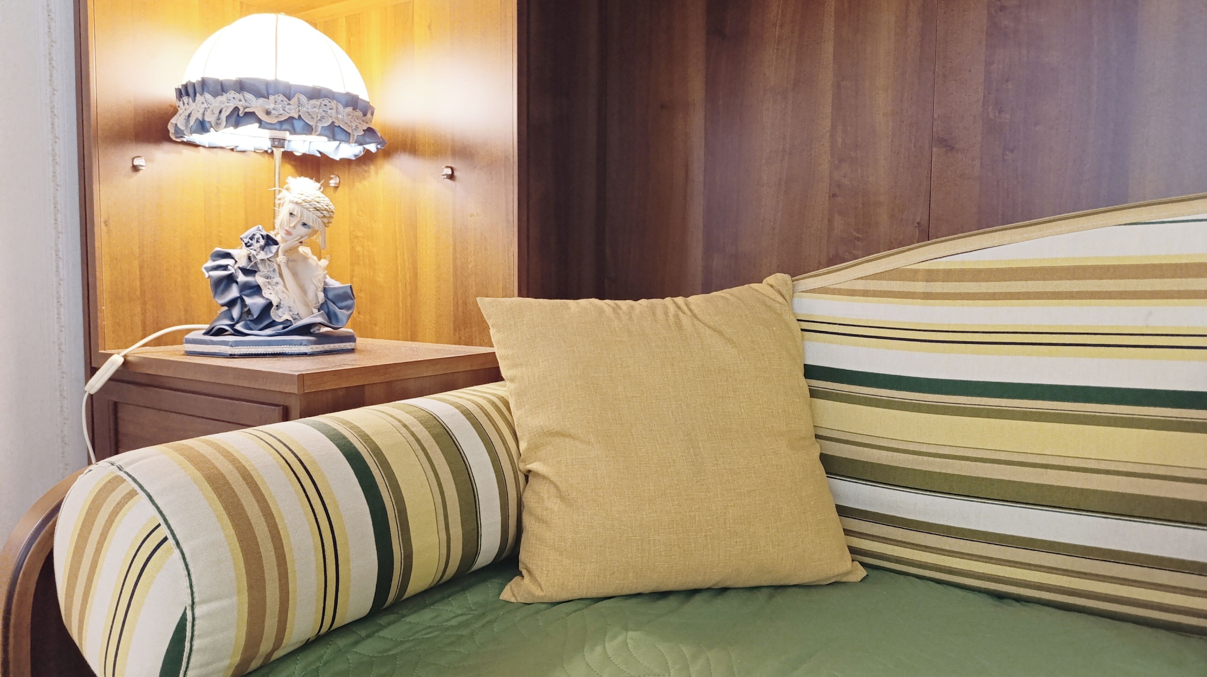 Un lit avec une literie à rayures, un oreiller beige et une lampe avec un abat-jour décoratif posée sur une table de chevet en bois.