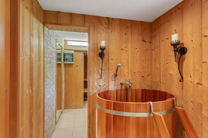 Un intérieur en bois avec une grande baignoire en bois, deux appliques murales et un tableau électrique apparent.