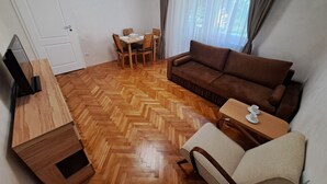A living room with a herringbone wooden floor, a brown sofa, a TV stand, a dining table with chairs, and a window with curtains. - Family Heritage from past to the future (Sibiu)