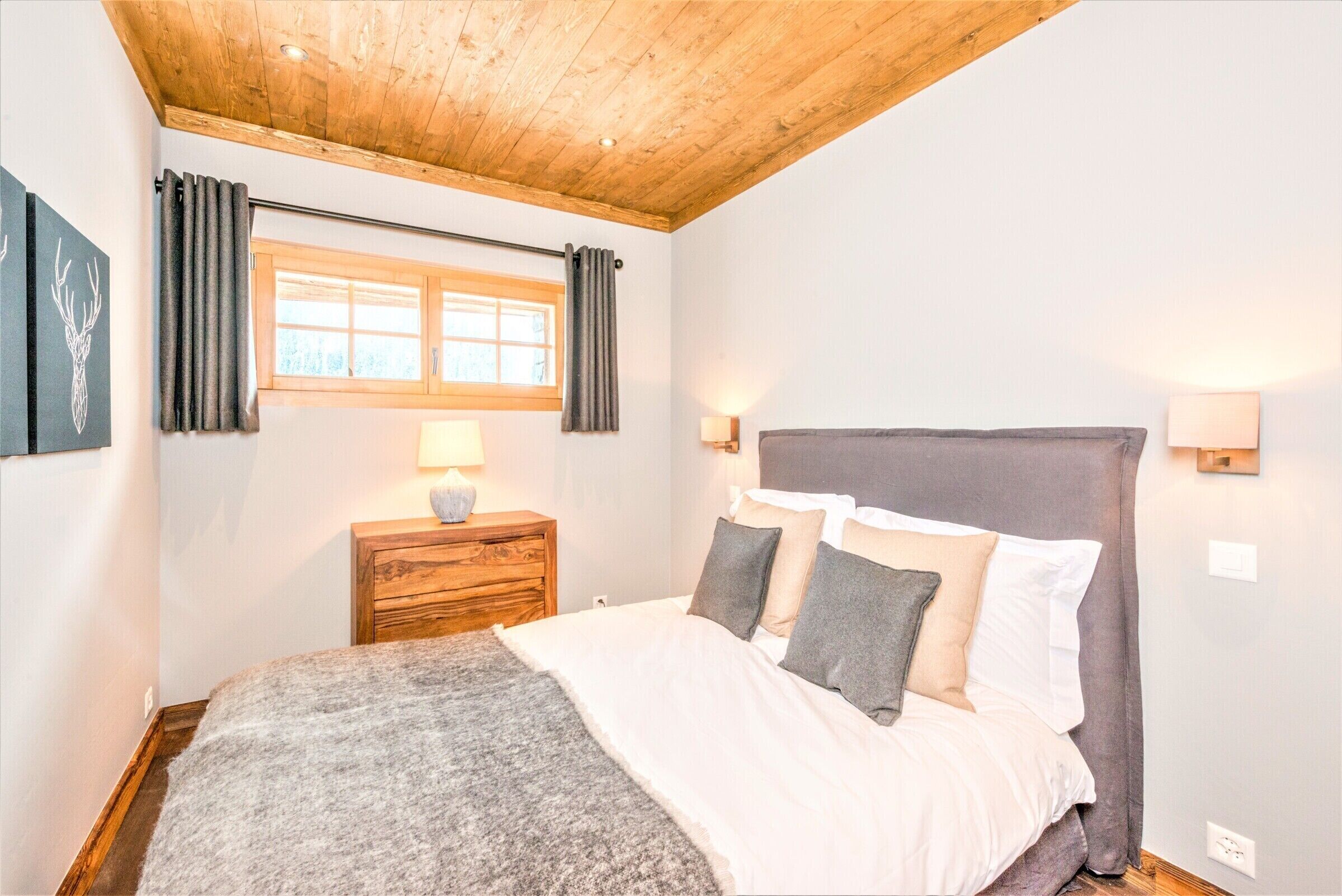 Un dormitorio con techo de madera, una cama con ropa de cama gris y blanca, una mesita de noche con una lámpara y una ventana con cortinas.