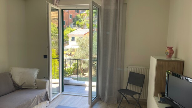 A modern living room with a sofa, a TV, a small table, and a chair. A sliding glass door leads to an outdoor balcony with a view of buildings and trees. - Oleandri cosy Apartment Near best beaches (Monterosso al Mare)