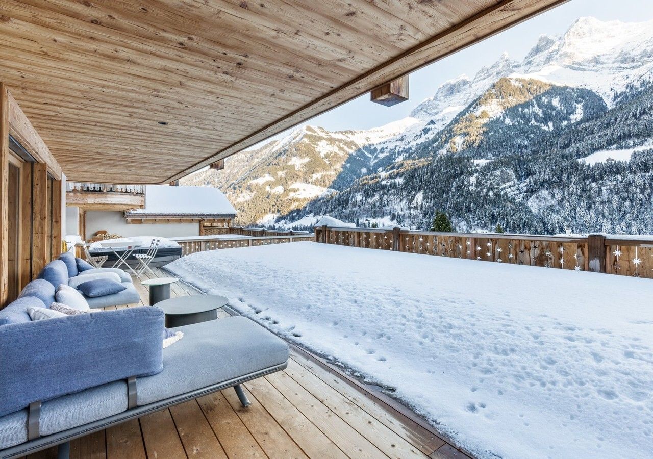 A wooden deck with a sofa and tables, overlooking a snowy mountain landscape.