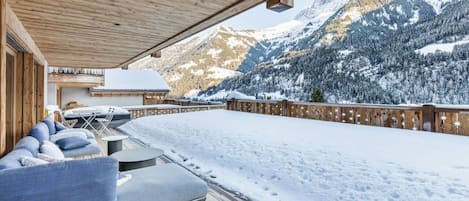 A wooden deck with a sofa and tables, overlooking a snowy mountain landscape.