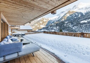 A wooden deck with a sofa and tables, overlooking a snowy mountain landscape.