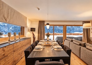 A dining area with a wooden table set for a meal, a sofa, and a view of snowy mountains through large windows.