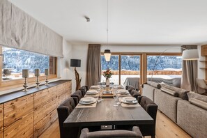 Uma sala de jantar com mesa de madeira posta para quatro pessoas, um sofá e vista para uma paisagem nevada.