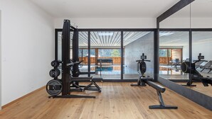 Une salle de sport moderne équipée d’une zone d’entraînement avec des poids, d’un rameur et d’un banc de développé couché. De grandes fenêtres offrent une vue sur une terrasse en bois et une piscine.