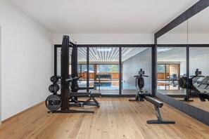 Et moderne fitnessrum med en vægttræningsstation, en romaskine og en bænkpres. Store vinduer giver udsigt til en træterrasse og en swimmingpool.