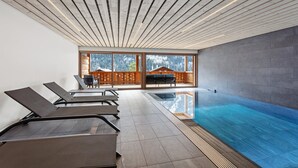 Une piscine intérieure moderne avec un plafond en bois, un espace détente avec des fauteuils et une vue sur un paysage montagneux grâce à de grandes fenêtres.