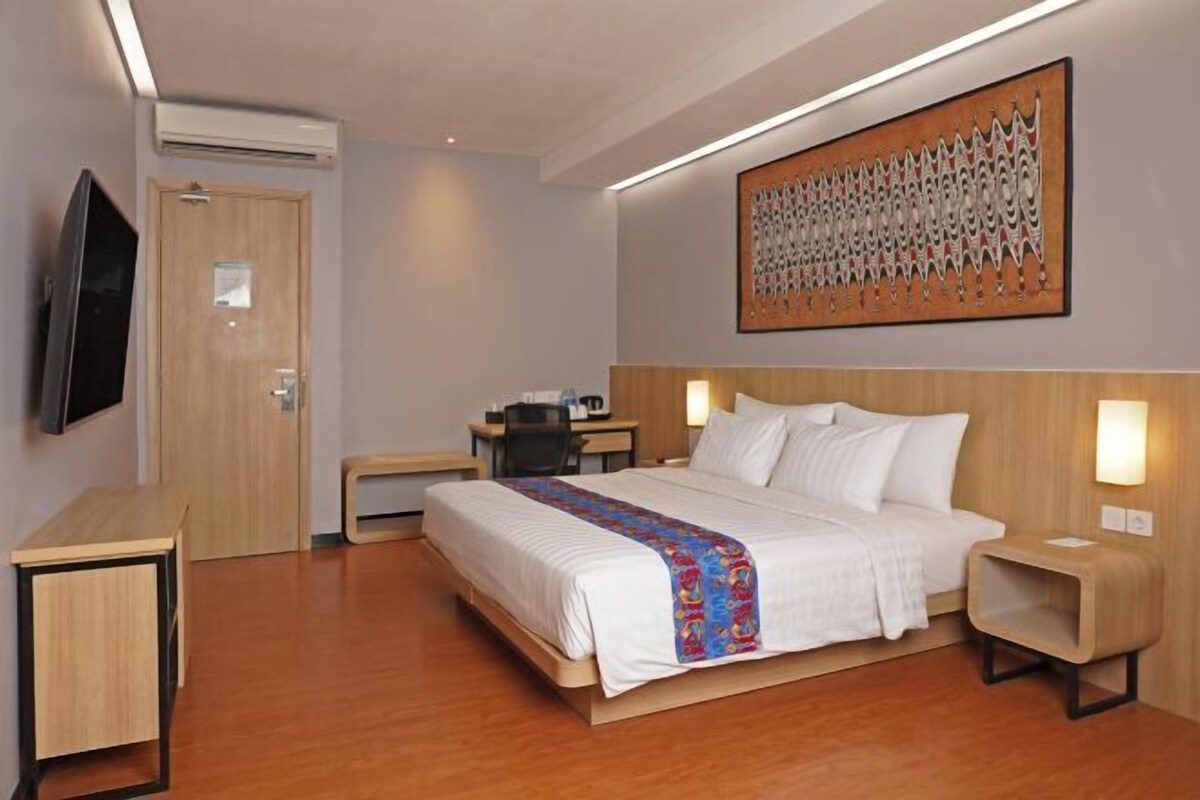 A hotel room with a bed, bedside tables, a desk, a chair, a TV, and a large abstract painting on the wall.