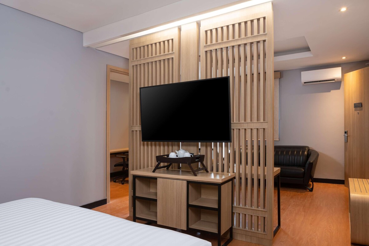 Quarto de hotel com televisão de tela plana fixada em parede de painel de madeira, cama com colchão branco, sofá de couro preto e escrivaninha de madeira com bandeja contendo um bule e xícaras.