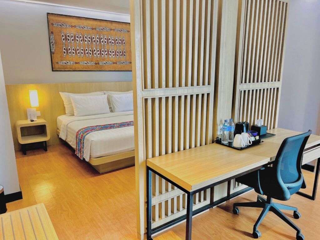A hotel room with a bed, bedside table, desk, chair, and a decorative wall panel.