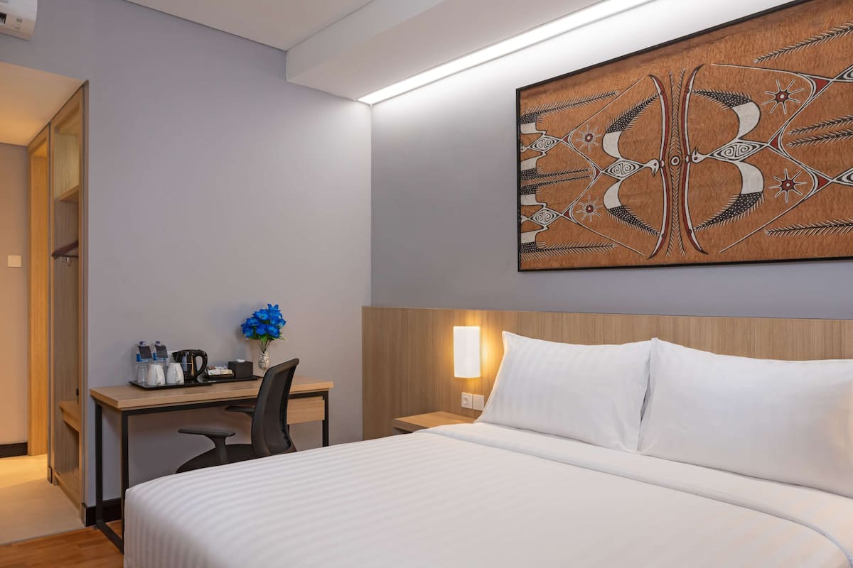 Ein Hotelzimmer mit einem Bett, einem Schreibtisch, einem Sessel, einem Nachttisch und einem an der Wand hängenden Kunstwerk.